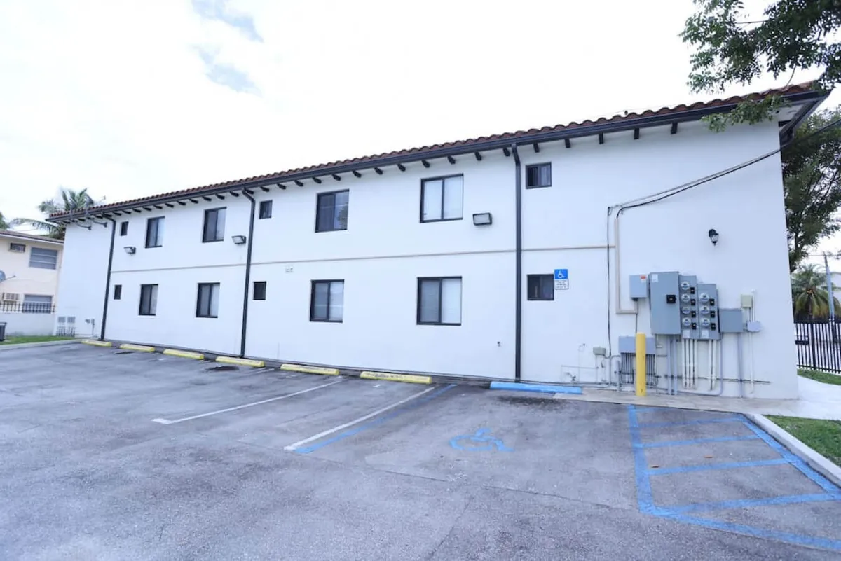 This exterior shot captures the building’s facade, windows, and a clearly visible parking area directly adjacent to the entrance. The image shows a well-maintained, accessible entry point, suggesting an easy transition from parking to the apartment.
