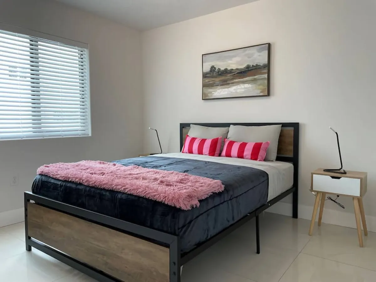 The image shows a serene bedroom with a cozy bed dressed in contrasting bedding and pillows, flanked by modern bedside tables and lamps. Above the bed hangs a vibrant landscape painting, adding color and personality to the restful space. The atmosphere is calm and inviting, perfect for unwinding after a day exploring Miami.