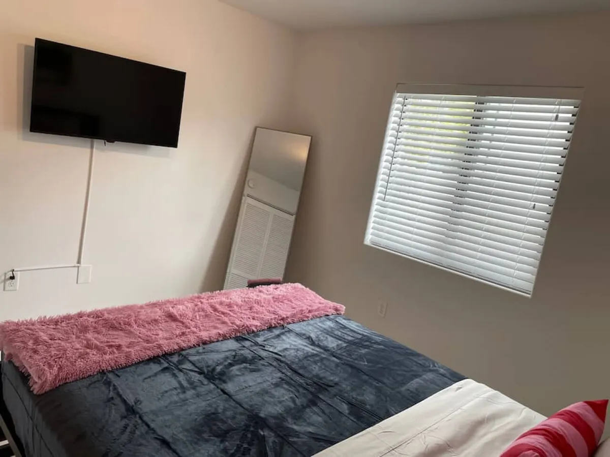 A cozy bedroom highlighted by a bed with colorful blankets and pillows. The room includes a wall-mounted flat screen TV, a floor mirror leaning against the wall, and a window with white blinds. The ambiance is calm, with elements for both relaxation and practical use, such as dressing and entertainment.