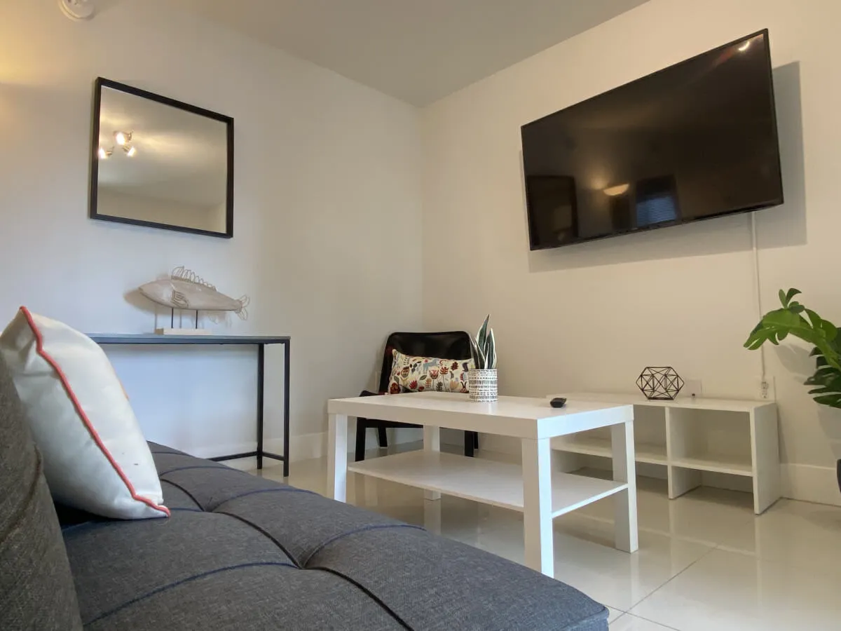 An inviting interior view with a large wall-mounted smart TV, stylish white coffee table, and a black chair with a patterned pillow. Decorative touches like a fish sculpture on a console table add personality. The setting is modern and calm, hinting at private, comfortable living accessible directly from outside.