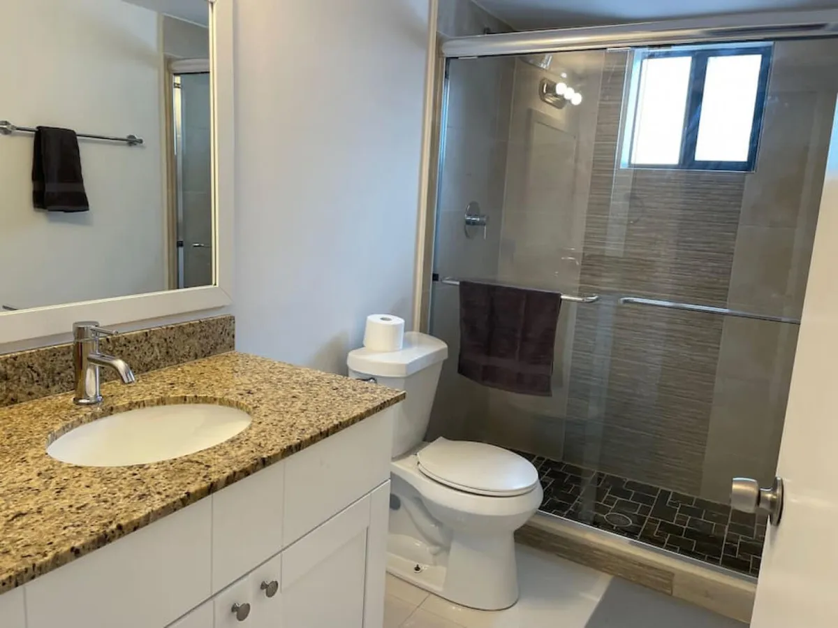 A sparkling full bathroom equipped with a granite countertop and sink, a toilet with a mounted roll, and a glass-enclosed shower with tiled interior. A large mirror sits above the sink, creating a fresh and inviting atmosphere ideal for morning and evening routines.