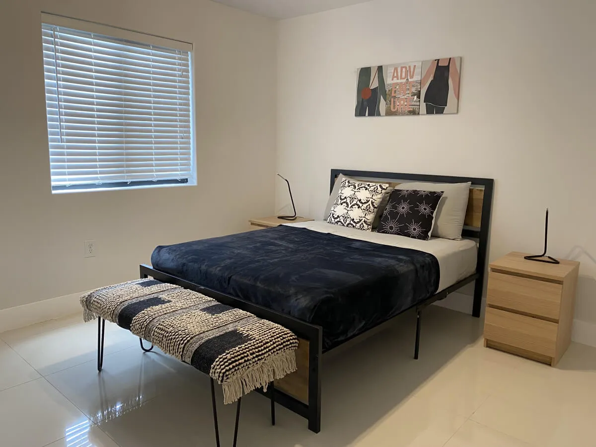 A tranquil bedroom featuring a modern bed adorned with dark bedding and decorative pillows. At the foot of the bed sits a woven bench, while simple wooden nightstands with minimalist lamps flank each side. Abstract wall art hangs above, and the overall setting is uncluttered and filled with gentle, natural light.