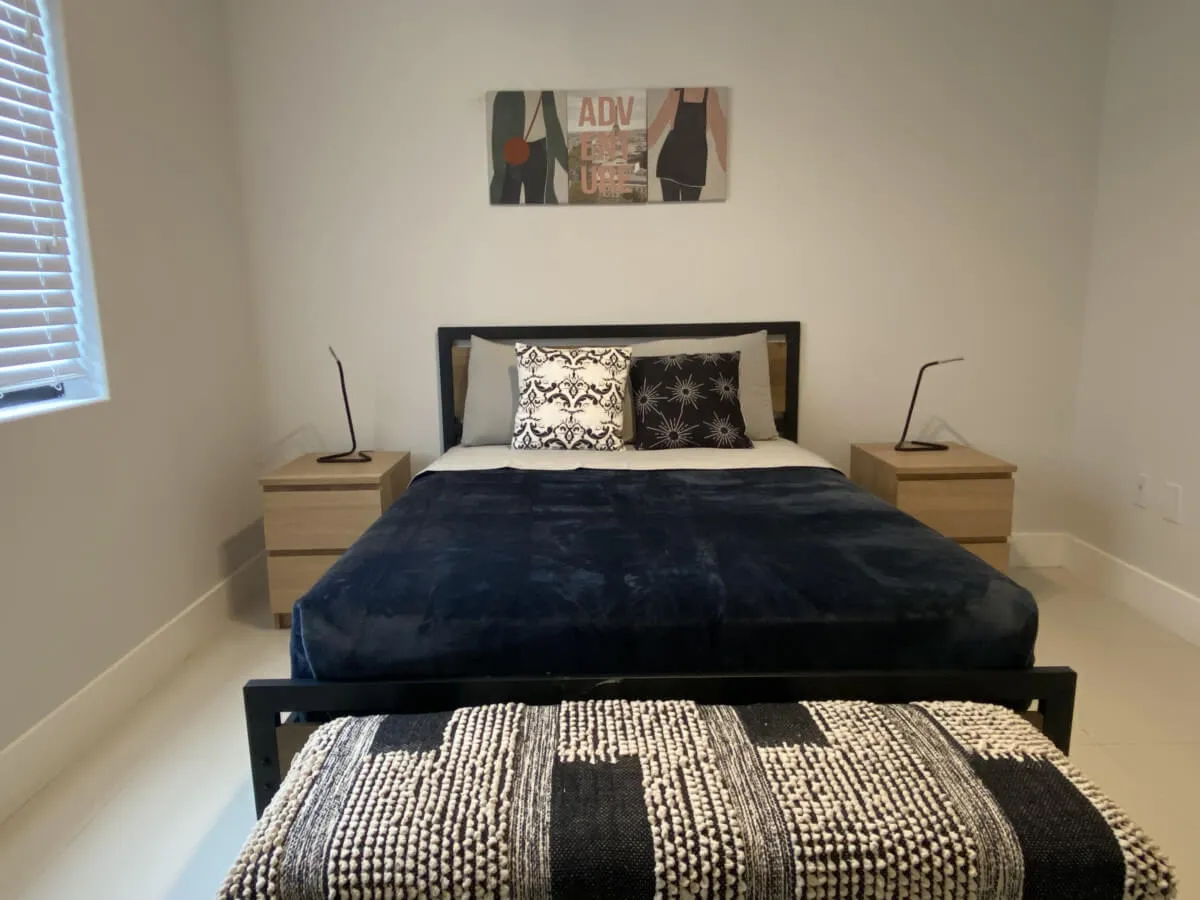 A serene bedroom scene with a bed dressed in a dark blue blanket and decorative pillows, flanked by wooden nightstands with modern lamps. A decorative bench sits at the foot of the bed, and stylish wall art hangs above.