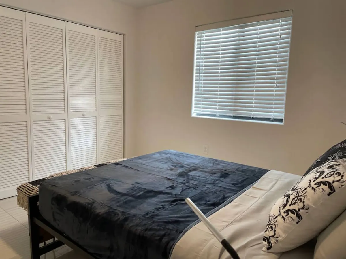 A bedroom featuring a bed with dark bedding and patterned pillows, a window with horizontal blinds, and white closet doors. The ambiance is restful and quiet, suggesting a comfortable private retreat after a busy day in Miami.