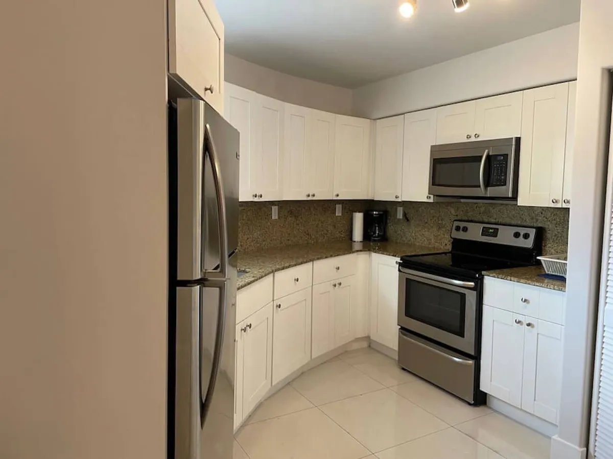 This image presents a bright, modern kitchen featuring a stainless steel refrigerator, oven with stove and microwave, granite countertops, and crisp white cabinetry. The setup feels welcoming and practical, ideal for preparing meals or morning coffee.