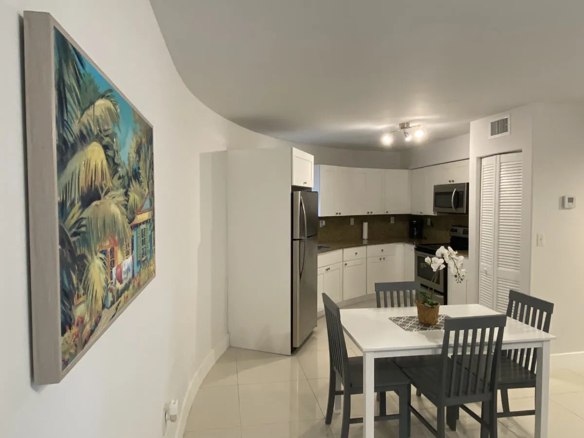 A cozy corner with a small dining table for four, surrounded by white kitchen cabinets and modern stainless steel appliances. A decorative plant on the table and a colorful tropical painting enhance the inviting atmosphere, making the area suitable for relaxed group gatherings.