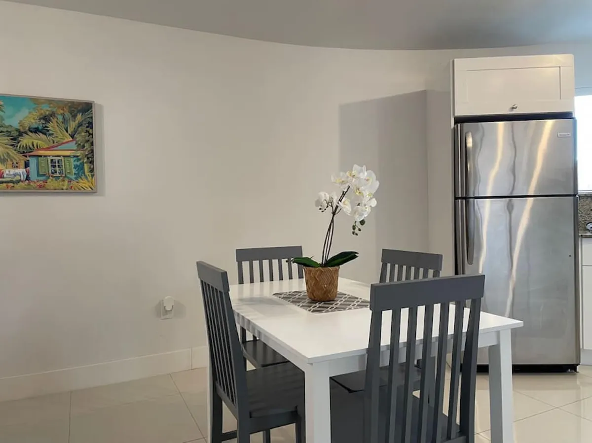 A bright, inviting dining area featuring a white table with gray chairs and a potted white orchid as a centerpiece. The space includes a stainless steel refrigerator and a colorful wall painting, suggesting a seamless connection to the kitchen. The overall atmosphere is open and welcoming, ideal for sharing meals and conversations.