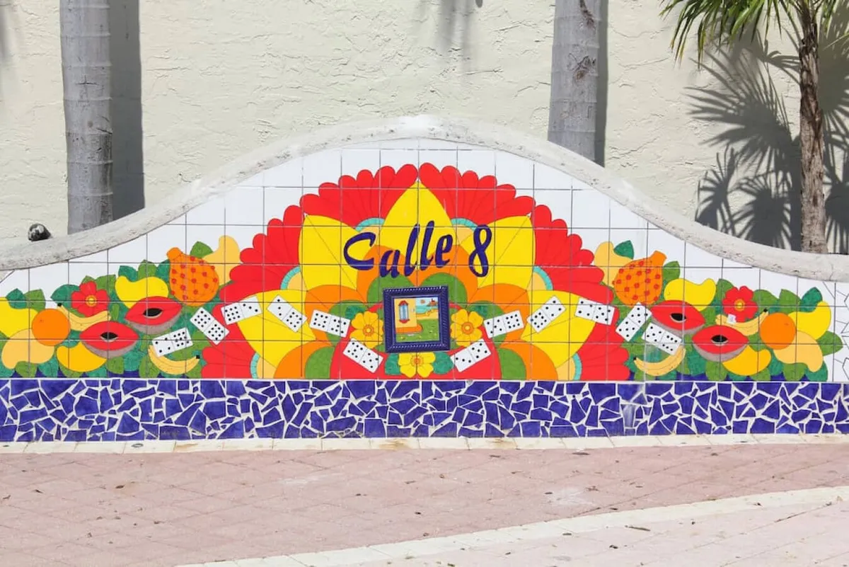A vibrant exterior mural with 'Calle 8' text, surrounded by colorful floral and fruit motifs, domino tiles, and palm trees. The scene captures the energetic and festive spirit of Miami's iconic Cuban district.