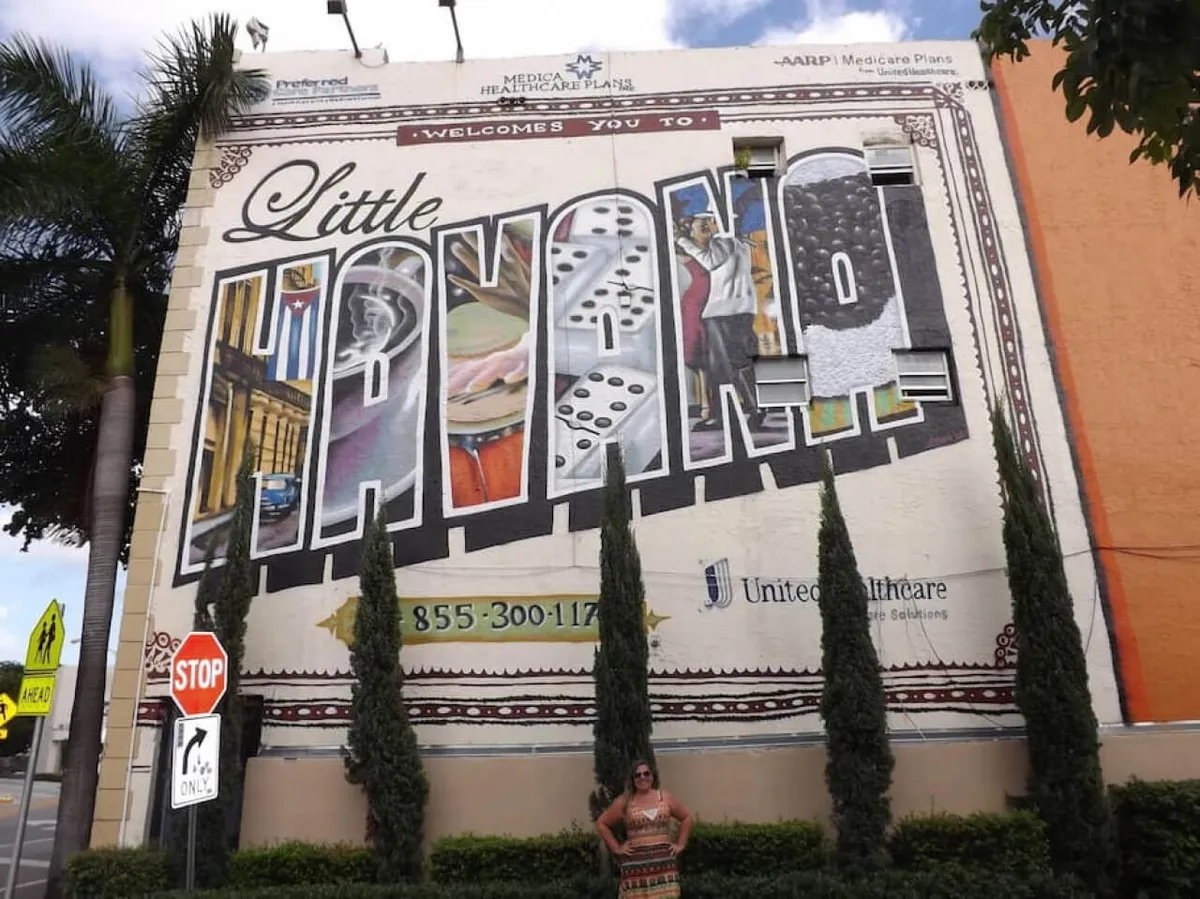 A vibrant exterior scene featuring the iconic 'Little Havana' mural. The mural is adorned with colorful Cuban cultural imagery and is set along a lively street with cypress trees, street signage, and a person enjoying the view. The area exudes a festive, welcoming atmosphere that captures the essence of Calle Ocho.