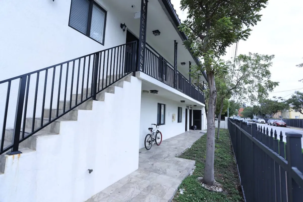 This exterior photo shows the white apartment building facade with a black metal railing and a secure black metal fence bordering the property. There is a concrete walkway leading directly to the door at ground level—no stairs required. A bicycle is parked near the entrance, and the overall scene feels quiet, safe, and welcoming, emphasizing both the ease of access and the secure, fenced environment.